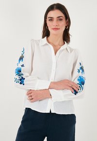 Chemise en lin blanc à manches longues avec un broderie florale bleue sur les poignets. Design à boutonnage avec une coupe décontractée.
