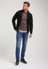 Giacca nera con zip sopra un maglione borgogna a consistenza, abbinata a jeans blu e sneaker scure. L'uomo si trova in posizione casual con una mano sui fianchi.