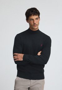 Svart polotröja i ett slätt stickat tyg. Har en liten röd logotyp på vänster bröst samt ribbstickade muddar och fåll.