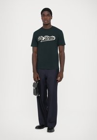 Melnas kokvilnas T-kreklu ar baltu tekstu, kas lasāms "The Kooples", apvienots ar tumšām svītrainām biksēm un melnu somu rokās.