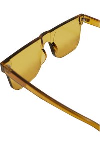 Ochelari de soare de culoare maronie, cu un design unghiular, lentile transparente galbene și o ramă îndrăzneață ce combină finisaje mate și lucioase.