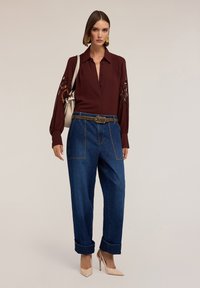 Blusa burdeos con mangas bordadas, combinada con pantalones vaqueros ajustados de tiro alto en azul con detalles de tachuelas; tacones altos nude.