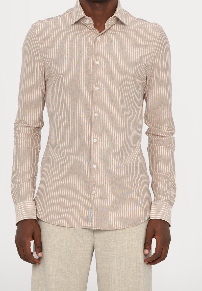 Homme portant une chemise beige à manches longues boutonnée avec des rayures verticales blanches et un pantalon beige clair sur un fond uni.