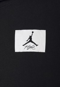 Tela negra con una etiqueta de ropa que presenta un fondo blanco, un logo de Jumpman en negro y la palabra "Flight" en cursiva.