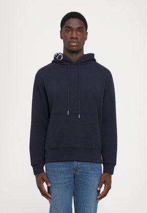 Giovane uomo che indossa un hoodie blu con tasca frontale e jeans blu, in piedi contro uno sfondo bianco uniforme.