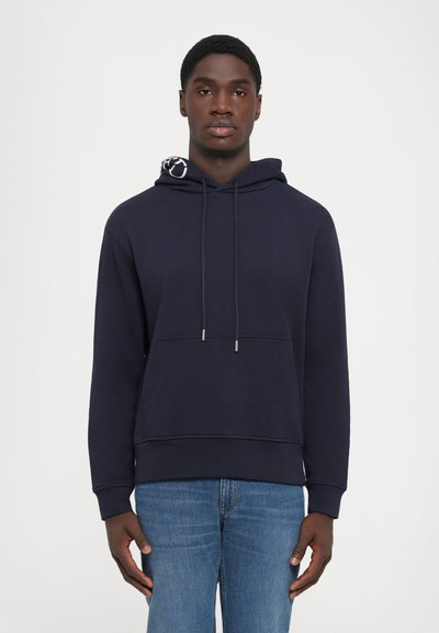 Joven con una sudadera azul marina con bolsillo frontal y jeans azules, de pie contra un fondo blanco liso.