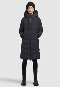 Doudoune noire avec un motif en chevrons, capuche et fermeture éclair, associée à un legging noir et des bottes hautes, sur un fond neutre.