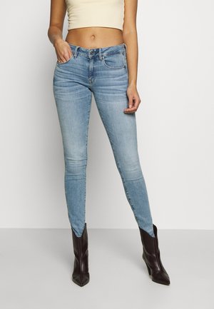 Slim fit lyseblå jeans med forlommer båret med midt-kalv sorte læderstøvler og en beige cropped top, stående mod en hvid baggrund.