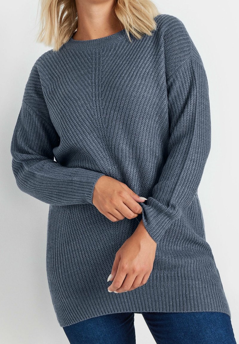 Suéter de punto gris con patrón texturizado, cuello redondo y mangas largas, superpuesto sobre denim azul. Presenta puños y dobladillo acanalados.