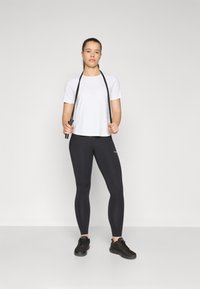 Maglietta atletica bianca a maniche corte, leggings neri e sneakers neri. Il modello tiene una corda per saltare nera, in piedi su uno sfondo neutro.