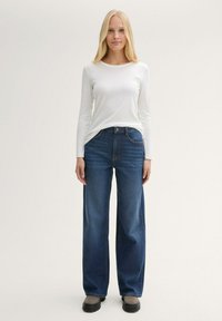 Bijela majica dugih rukava u kombinaciji s plavim širokim farmerkama. Ovaj outfit ima glatku teksturu i klasičan denim izgled, a upotpunjuju ga sivi cipele.