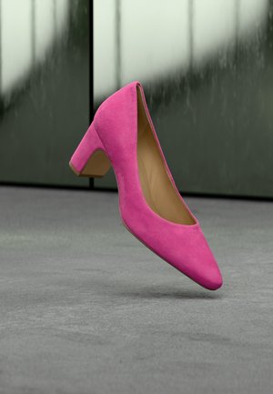 Felroze suède midhakkenpomp zwevend boven een grijze vloer met een wazige groen-grijze muur op de achtergrond.