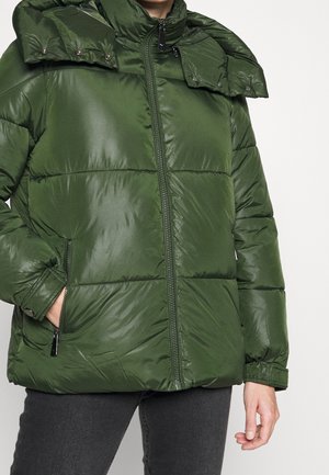 Zielona pikowana kurtka z przednim zamkiem błyskawicznym, odpinanym kapturem na zatrzaski, kieszeniami na zamek oraz elastycznymi mankietami, noszona przez osobę w ciemnych spodniach.