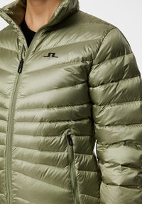 Vihreä pehmustettu takki vaakasuorilla saumoilla. Varustettu korkealla kauluksella, mustalla vetoketjulla ja huomaamattomalla logolla vasemmassa rinnassa.