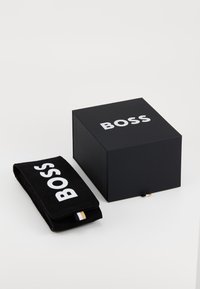 Μαύρο κουτί δώρου με λευκό λογότυπο "BOSS" στην κορυφή, συνοδευόμενο από ένα αντίστοιχο μαύρο σουέτ τσαντάκι με το ίδιο λογότυπο και μια λεπτομέρεια με λωρίδα στο πλάι.