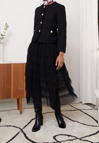 Černá texturová bunda se zlatými knoflíky, která je spojena s černou vrstvenou tylovou sukní. Modelka má na sobě černé kozačky na vysokém podpatku do kolen.