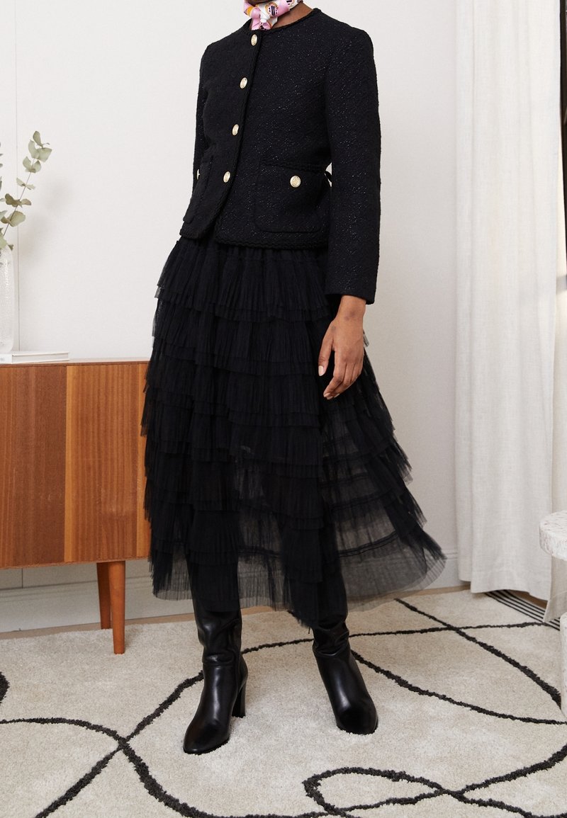 Černá texturová bunda se zlatými knoflíky, která je spojena s černou vrstvenou tylovou sukní. Modelka má na sobě černé kozačky na vysokém podpatku do kolen.
