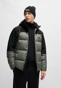 Padded jacket in olive green and black with a zip closure, two-tone design, and logo on the chest. Worn with a black beanie.