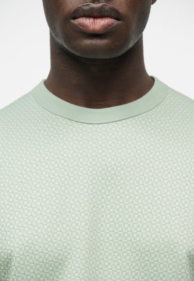 Εμφάνιση κοντινού πλάνου του κάτω μέρους του προσώπου και του λαιμού ενός ατόμου που φοράει ανοιχτοπράσινο πουκάμισο με μοτίβο και στρογγυλή λαιμόκοψη, μπροστά σε απλό φόντο.