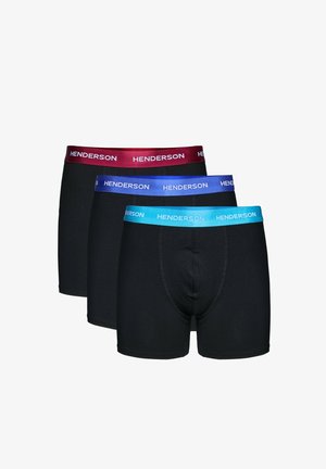 Hemdloser Mann mit kurzen Haaren trägt schwarze Boxer-Shorts mit rotem Bund und der Aufschrift "Henderson" und steht vor einem schlichten hellen Hintergrund.