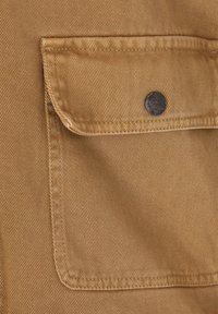 Nærbillede af tan-stof, der viser en lomme med en metalknap, med en lige tekstur og synlige sømdetaljer.