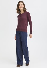 Bordeauxfarbener Cardigan mit Rundhalsausschnitt und Knopfschluss, kombiniert mit weiter geschnittenen dunkelblauen Jeans und braunen Schuhen.