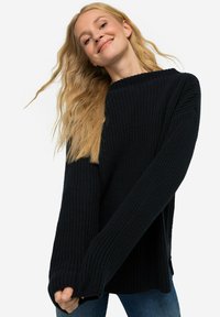 Schwarzer übergroßer Strickpullover mit gerippter Textur und langen Ärmeln, ausgestattet mit seitlichen Schlitzen und einem weit ausgeschnittenen Hals, kombiniert mit blauen Jeans.