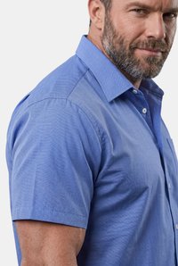 Mand med trimmet skæg iført en kortærmet blå skjorte med knapper, vendt fremad og kigger let til sin venstre side.