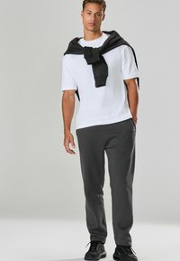 Grijze joggingbroek gecombineerd met een wit T-shirt, een zwart sweatshirt om de schouders gedrapeerd, en zwarte sportschoenen. Eenvoudig ontwerp, casual pasvorm.