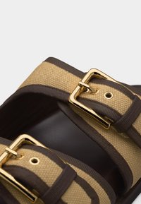 Κοντινό πλάνο από μπεζ και καφέ υφαντικές λουριά σανδαλιών με χρυσά μεταλλικά αγκράφες σε σκουρόχρωμη σόλα.