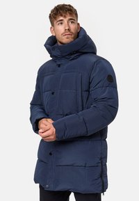 Doudoune bleu marine avec col haut, fermeture à boutons et poches zippées. Offre un tissu texturé et un design matelassé.