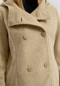 Beige topelt rinnaga mantel tekstuurses kangas, suur krae ja kaks eesmist taskut. Omab viit ümmargust nuppu ja kapuutsi.