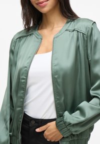 Giacca bomber in satin verde con design senza colletto, polsini elasticizzati e zip frontale. Indossata sopra una maglietta bianca e pantaloni neri.