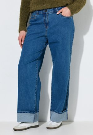 Person iført blå vidde jeans med opbukket kant, hvide sneakers og en grøn fuzzy sweater, stående med den ene hånd i lommen.