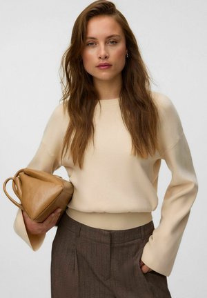 Kvinde med langt brunt hår iført en cremefarvet sweater og brune bukser, der holder en lysebrun lædertaske mod en ensfarvet baggrund.