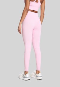 Kobieta w różowych legginsach z wysokim stanem i pasującym krojem topu, stojąca w białych sneakersach na jednolitym jasnoszarym tle.