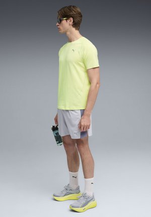 Leuchtend gelbes sportliches T-Shirt mit kurzen Ärmeln, kombiniert mit grauen Shorts und grauen Sneakers mit gelben Akzenten. Hält eine transparente Wasserflasche.