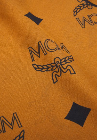 Oransje stoff med et gjentakende navy-blått MCM-logo, dekorativ krans og firkantede detaljer. Materialet ser glatt ut med en lett tekstur.