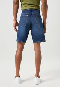 Granatowe szorty dżinsowe o prostym kroju, widoczne szwy i dwie tylne kieszenie z wyraźnym wzorem. Noszone z białymi sneakersami.