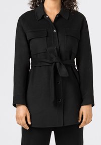 Svart, långärmad skjortjacka med två bröstfickor, ett bälte i midjan och knäppning fram. Mjuk, texturerad tyg.