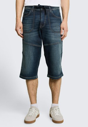 Mann, der dunkelblaue Denim-Knielange Shorts mit Fronttaschen, Kordelzug in der Taille, weiße Knöchelsocken und beige Turnschuhe trägt.