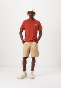 Red cotton T-shirt with logo, beige cotton shorts, and light-colored sneakers. Model wearing a white bucket hat, hands in pockets.