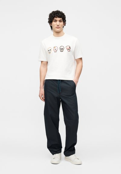 Νέος άνδρας με σγουρά μαλλιά που φοράει λευκά αθλητικά παπούτσια, άνετο μαύρο παντελόνι και λευκό μπλουζάκι με τέσσερα πολύχρωμα σχέδια κρανίων.