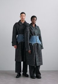 Dos modelos visten abrigos oscuros de gran tamaño en gris con grandes cuellos y cinturones de mezclilla azul. Los abrigos tienen un acabado texturizado y mangas largas.