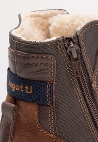 Dunkelbrauner Knöchelstiefel aus strukturiertem Stoff, mit Lederakzenten und einem Reißverschluss. Gefüttert mit weichem Kunstfell, mit einem blauen Stofflogo-Riemen.