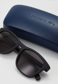 Ochelari de soare negri cu finisaj mat, așezați lângă o carcasă albastră Lacoste cu logo embosat, evidențiind designul elegant și funcționalitatea.