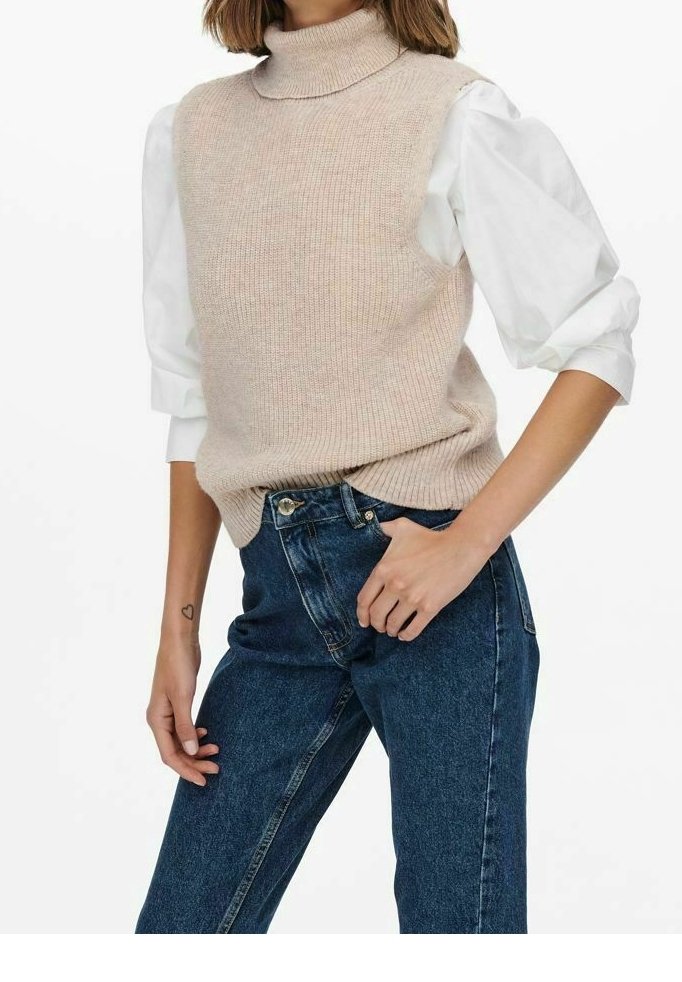 Beige gebreid coltrui-vest met geribbelde textuur, gedragen over een witte blouse met opgeblazen mouwen. Gecombineerd met een hoge taille blauwe spijkerbroek.