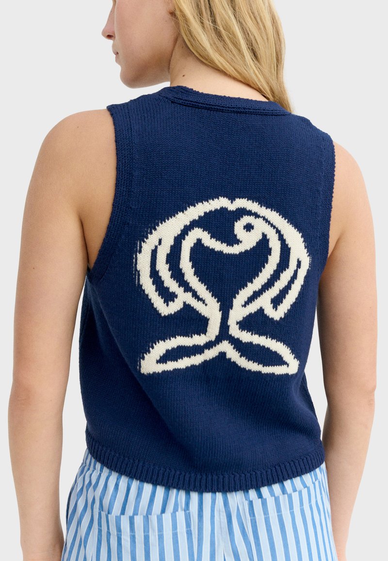 Personne blonde portant un gilet sans manches en maille bleu marine avec un grand motif abstrait en forme de cœur blanc dans le dos.