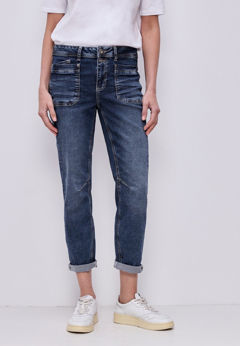 Jean denim bleu foncé slim ajusté retroussé, porté avec des baskets blanches et une chemise blanche à manches courtes sur un fond uni.