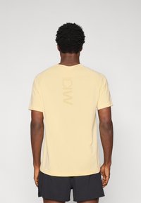 Φωτεινό κίτρινο αθλητικό μπλουζάκι με κοντά μανίκια ράγκλαν, με κάθετη εκτύπωση λογότυπου στην πλάτη και λείο, ελαφρύ ύφασμα.